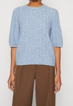 Personne portant un pull côtelé bleu clair avec des manches aux coudes et un pantalon marron ample taille haute, debout devant un fond uni.