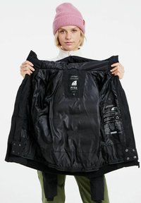 Schwarze, isolierte Jacke mit einem glänzenden Innenfutter. Hat ein Etikett mit der Aufschrift "PYRE Green" und verstellbare Druckknöpfe am Saum. Getragen von einer Person mit einer pinken Mütze.
