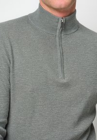Grauer, strukturierten Zip-Hals-Pullover mit hohem Kragen und glattem Reißverschluss, der ein modernes Design und vielseitigen Stil zeigt.