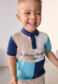 Stickad kortärmad polo för barn i beige, marinblå och turkos block, med broderad text "Les Petits Champions Club."