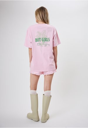 Kobieta w jasnoróżowej, oversize koszulce z napisem i grafiką "HOT GIRLS grow their own veggies", różowych szortach, białych skarpetkach i beżowych botkach.