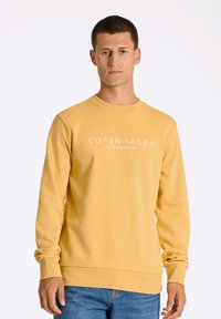 Gul sweatshirt av mjukt tyg med rund halsringning och ärmar med mudd. Framsidan har texten "COPENHAGEN LINDBERGH" i vitt.
