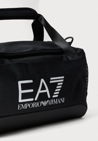Borsa da viaggio in tela nera con chiusura a zip, manico imbottito e logo EA7 Emporio Armani bianco. Presenta una finitura testurizzata e un pannello laterale in rete.