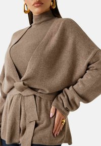 Cardigan beige lavorato a maglia con design incrociato, maniche lunghe e cintura annodata in vita. Presenta un sottostrato con collo alto. Texture liscia.