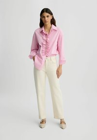 Chemise rayée rose et blanche avec un détail froufrou à l'avant, associée à un pantalon droit crème et des chaussures beiges ornées.
