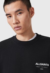 Young man with short hair wearing a black ALLSAINTS sweatshirt with white Japanese and English text on a light background.