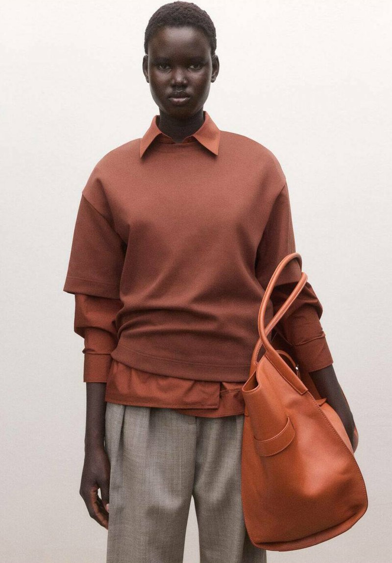 Femme portant des hauts superposés de couleur rouille et un pantalon gris, tenant un grand sac à main en cuir rouille avec double poignée.