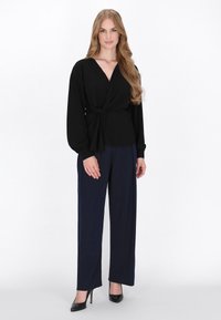 Zwarte wikkelblouse met een diepe V-hals, lange mouwen en een strik in de taille, gecombineerd met marineblauwe straight-leg broek en zwarte hakken.