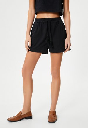 Schwarze, strukturierte Shorts mit einem elastischen Bund und Kordelzug, ausgestattet mit zwei Seitentaschen und Quastendetails; kombiniert mit braunen Slippern.