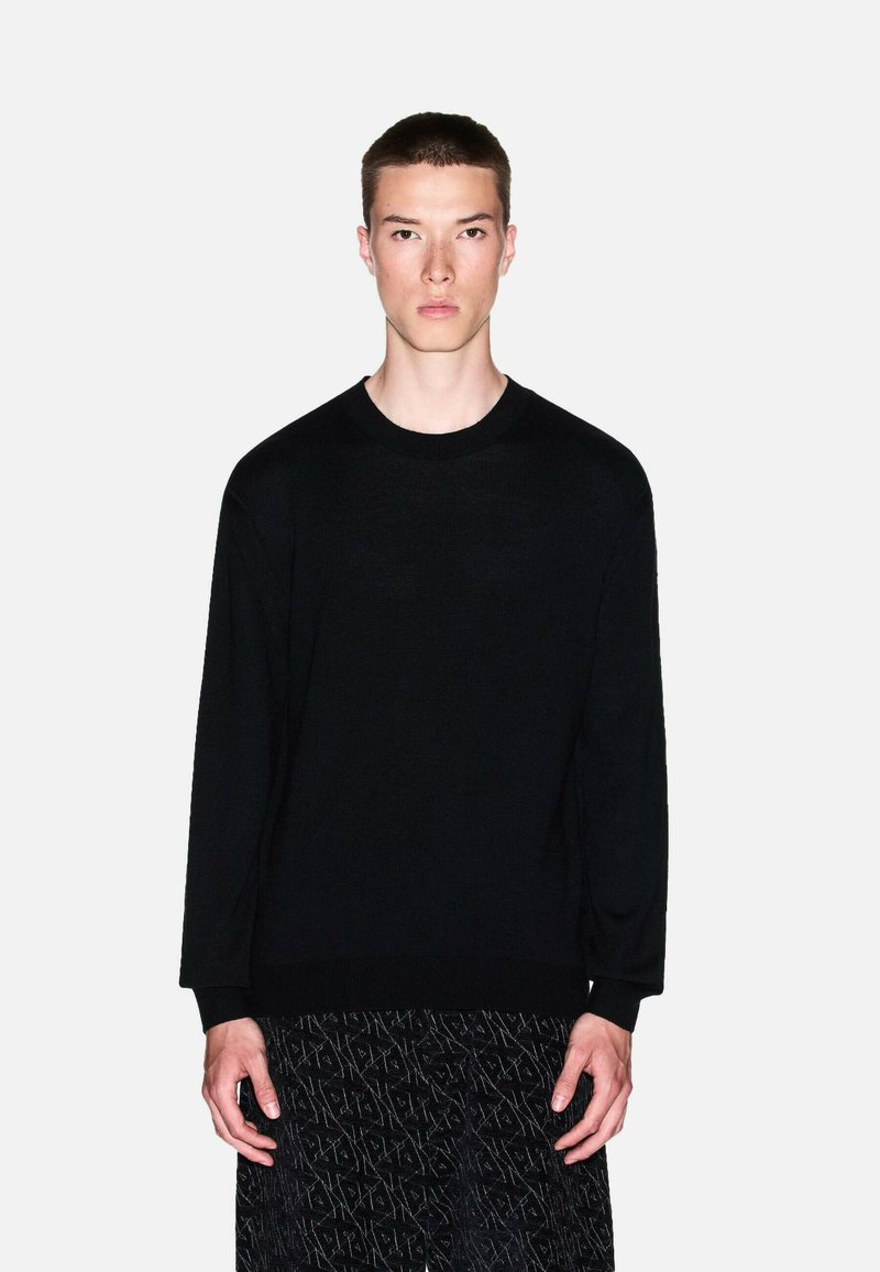 Maglione nero a maniche lunghe con scollo rotondo, texture lavorata a maglia e vestibilità comoda. Indossato sopra pantaloni scuri con fantasia, design minimale senza loghi.