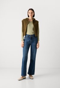 Strikket olivengrønn cropped vest over en lys grønn langermet topp, kombinert med blå flagrende jeans og beige flat sko.
