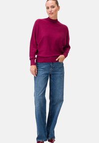 Fuchsia gestrickter Pullover mit hohem Kragen und lockerer Passform, kombiniert mit blauen Jeans im geraden Schnitt. Sanfte Textur, minimale Details.
