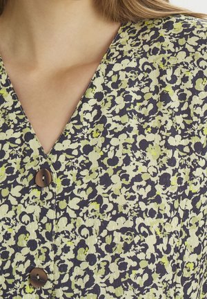 Nahaufnahme eines Ausschnitts mit einer dunklen Bluse mit gelb-grünen Blumen und zwei braunen Knöpfen vorne.