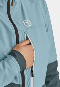 Light blue jacket with black zippers and gray cuffs; features a front pocket with a zipper. Logo displayed on the chest area.