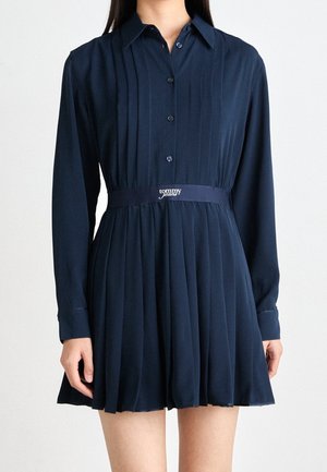 Nainen pukeutuneena tummansiniseen plissee-mekkoon, jossa pitkät hihat, napit edessä ja vyötäröllä "tommy jeans" -merkintä.