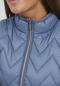 Ljust blå puffer-väst med hög krage, med ett chevron-quiltmönster och en silverdragkedja. Tillverkad av ett slätt, glänsande material.