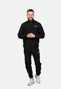 Schwarzer Trainingsanzug mit Reißverschlussjacke und Hose mit elastischem Bund. Auffälliges Logo auf Jacke und Hose, kombiniert mit weißen Turnschuhen.