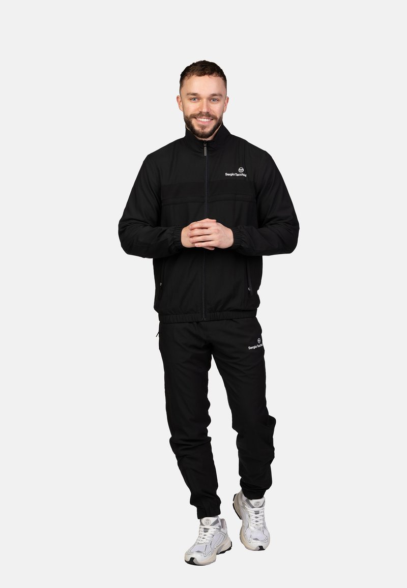 Schwarzer Trainingsanzug mit Reißverschlussjacke und Hose mit elastischem Bund. Auffälliges Logo auf Jacke und Hose, kombiniert mit weißen Turnschuhen.