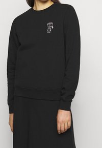 Svart sweatshirt gjord av bomullsblandning, med rund halsringning, långa ärmar och en liten broderad figur på bröstet. Mjuk textur.