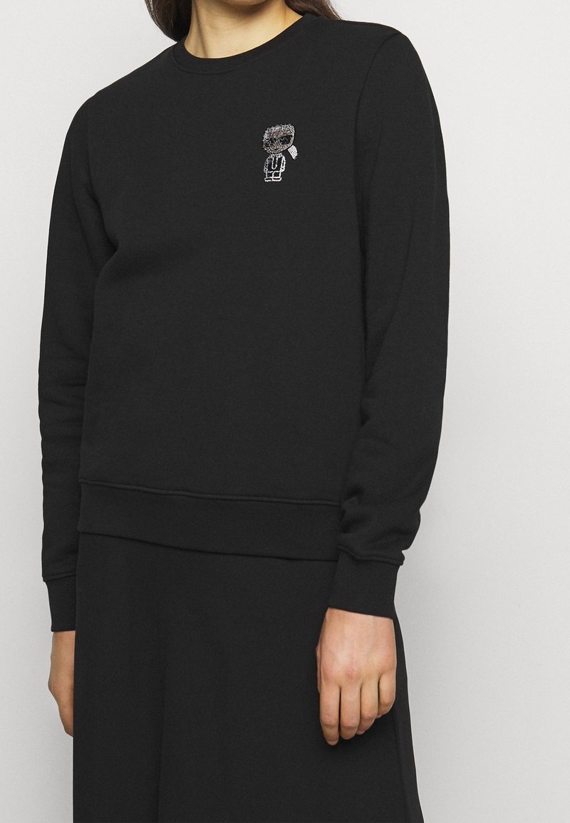 Svart sweatshirt gjord av bomullsblandning, med rund halsringning, långa ärmar och en liten broderad figur på bröstet. Mjuk textur.