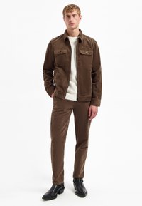 Bruine corduroyjas met twee borstzakken in combinatie met bijpassende bruine corduroypantalon. Model draagt een lichte trui en zwarte laarzen.