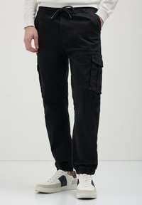 Mann in schwarzen Cargo-Hosen mit elastischen Knöcheln und weißen Sneakers mit schwarz-beigen Details, der vor einem schlichten hellen Hintergrund steht.