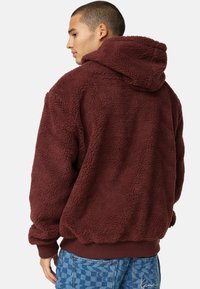 Felpa con cappuccio color bordeaux in pile, con una finitura testurizzata e soffice, un ampio cappuccio, polsini e orlo a costine. Presenta una vestibilità comoda e spalle scese.