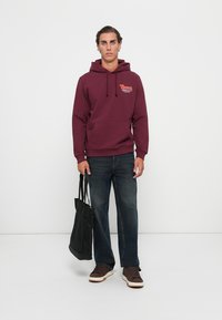 Mørkerød hoodie med forlommer, snoretræk og logo på brystet; parret med mørke jeans og brune sneakers; holder en sort tote bag.