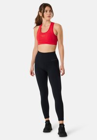 Brassière de sport rouge avec des bretelles larges, leggings taille haute noirs et chaussures de sport noires. Tissu lisse, design ajusté, minimalisme de la marque visible.