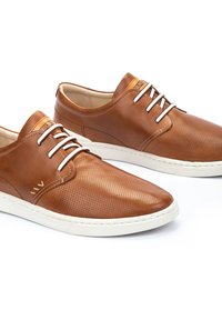 Zapatos de cuero marrón claro con textura perforada, suelas de goma blanca y cordones en contraste. Diseño de punta redondeada, estilo casual elegante, con acentos mínimos.
