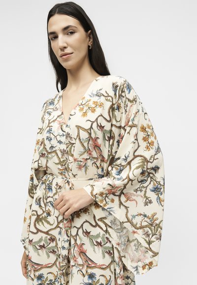 Γυναίκα με μακριά σκούρα μαλλιά που φοράει μία κρεμ ρόμπα σε στυλ κιμονό με λουλουδάτο σχέδιο και ζώνη στη μέση, σε απλό φόντο.