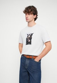 Άσπρο βαμβακερό μπλουζάκι με γραφικό ενός σκύλου που γαυγίζει, συνδυασμένο με μπλε τζιν παντελόνι και καφέ δερμάτινη ζώνη.