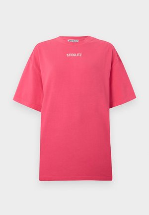 Persoon draagt een oversized roze T-shirt met de tekst "STIEGLITZ", losse grijze broek, zwarte schoenen en een zwarte baseballpet, staand tegen een witte achtergrond.