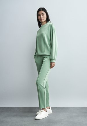 Γυναίκα όρθια που φοράει ανοιχτοπράσινο φούτερ με μακριά μανίκια και παντελόνι που ταιριάζει, μαζί με λευκά αθλητικά παπούτσια, μπροστά σε έναν μονόχρωμο γκρι τοίχο.