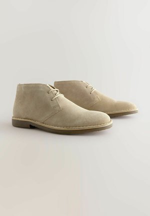 Beige Wildlederschuhe mit runden Zehen, Schnürung vorne und kontrastierenden braunen Gummisohlen. Glatte Textur mit subtilen Steppdetails.