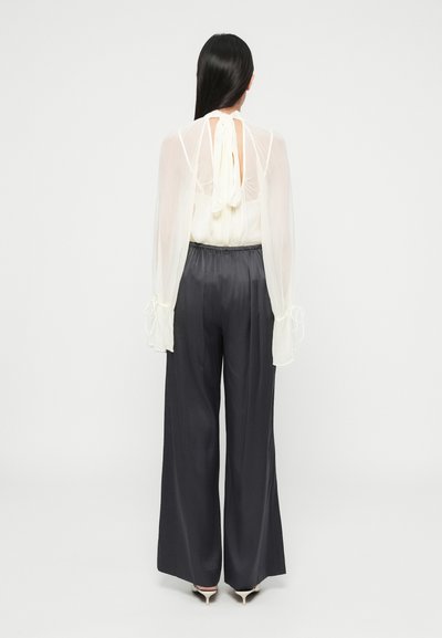 Blouse blanche transparente à manches fluides avec un détail noué dans le dos, associée à un pantalon large gris foncé et des talons clairs.