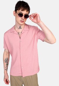 Camisa rosa de manga corta con un patrón texturizado, con cuello abotonado en la parte delantera y un ajuste relajado; combinada con gafas de sol redondas y pantalones de color claro.