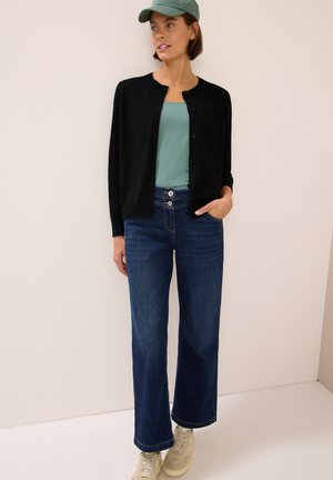 Zwarte cardigan met ronde hals, teal tanktop, wijde donkere denimjeans en lichtgekleurde sneakers. Pet in bijpassend teal kleur.