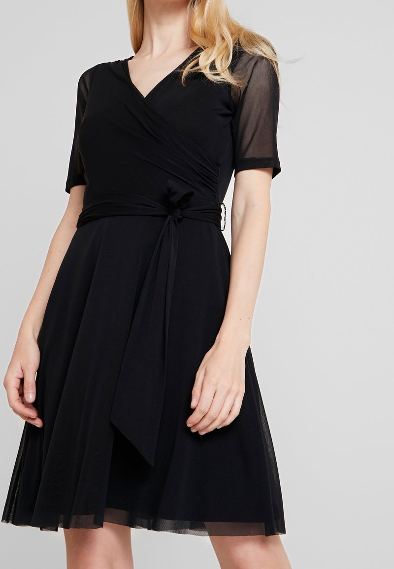 Femme portant une robe portefeuille noire avec des manches courtes en tissu transparent, une ceinture nouée à la taille et une jupe fluide longueur genou.