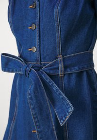 Denimmekko, jossa on tiukka malli, tummansinisestä kankaasta, jossa on viisi hopeista nappia ja sidottu vyötärövyö. Kontrastitikkaus näkyvissä.
