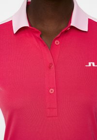 Rosa polo skjorta med vit krage, tre knappar och en vit logga på vänster sida. Slät tyg och figurnära design.