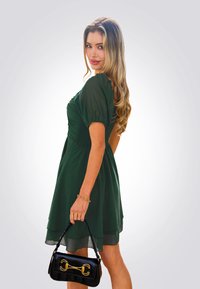 Vestido verde con mangas abullonadas, corpiño ajustado y falda fluida. Bolso negro con herrajes dorados. Tela suave, diseño en capas.