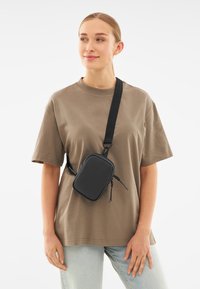 Borsa a tracolla rettangolare nera realizzata in materiale liscio, dotata di due zip e di una tracolla regolabile, indossata su una maglietta beige.