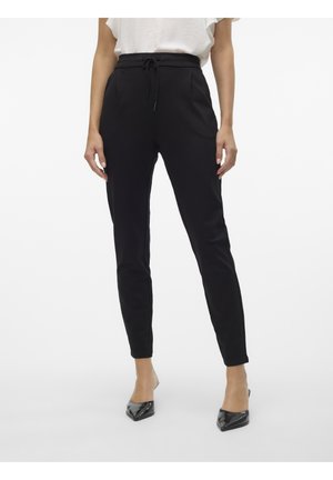 Femme portant un pantalon noir fuselé avec taille à cordon et des escarpins noirs à bout pointu, associée à un haut blanc à manches courtes.