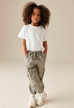 T-shirt bianco a maniche corte abbinato a pantaloni cargo verde oliva chiaro, caratterizzati da più tasche e polsini elastici, completato da sneakers bianche.