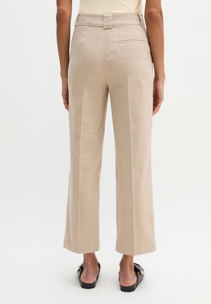 Achteraanzicht van een persoon die beige wijde cropped broek en zwarte instapschoenen draagt, staand tegen een effen achtergrond.