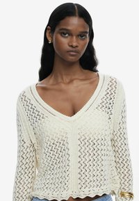Strickpullover in Creme mit V-Ausschnitt, langen Ärmeln und einem strukturierten Muster. Mit gewelltem Saum und detaillierter Webung im gesamten Design.