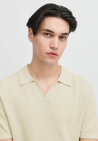 Beige kötött pólóing, texturált mintával, rövid ujjakkal és kivágott gallérral. Egyszerű, sokoldalú dizájn, amely alkalmas hétköznapi viseletre.