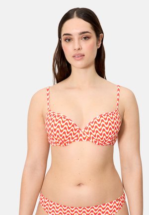 Mujer con cabello largo y castaño, vistiendo un sujetador estampado con zigzag rojo y blanco y pantalones de bikini a juego, sobre un fondo liso y claro.