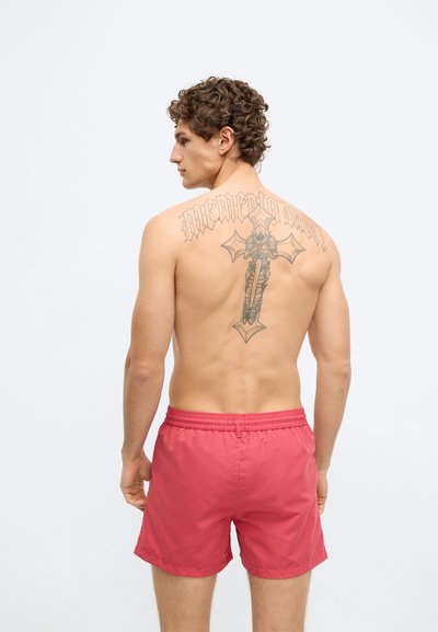 Röda badshorts med elastisk midjeresår, midjelånga, slät textur och en bakifrån vy som visar en stor, detaljerad tatuering.
